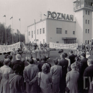 duża grupa ludzi zgromadzona na płycie lotniska, w głębi jasny budynek z wieżą i napisem Poznań