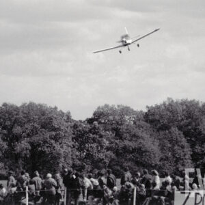 duża grupa widzów obserwuje niski przelot samolotu, w głębi drzewa