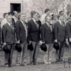 siedmiu mężczyzn stoi na baczność w jednym rzędzie, w prawej ręce trzymają kapelusze, za nimi grupa ludzi