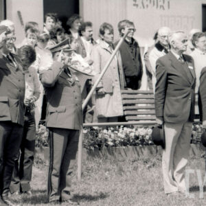 z lewej dwóch mężczyzn w mundurach, stoi i salutuje, z prawej, dwóch w garniturach stoi na baczność, w prawej dłoni trzymają kapelusze, z nimi grupa ludzi