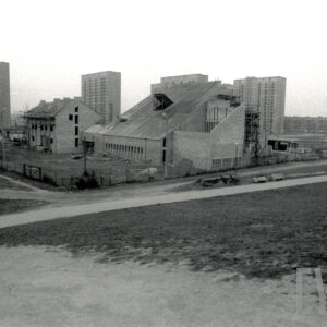 kompleks budynków sakralnych w trakcie budowy, na pierwszym planie budynek główny ze spadzistym dachem i malutkimi oknami z niższa przybudówką bez położonego jeszcze dachu, z nim budynek dwukondygnacyjny ze spadzistym dachem, w głębi wysokie bloki mieszkalne z wielkiej płyty