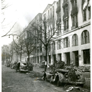 wypalone i zniszczone budynki wzdłuż ulicy, przed nimi gruzy, wzdłuż ulicy drzewa pozbawione liści i wypalone wraki samochodów