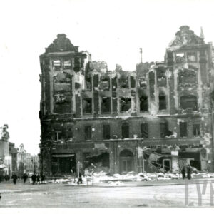 zniszczony i wypalony kilkukondygnacyjny budynek, przed budynkiem gruz leżący na chodniku, z lewej ulica idąca w dół, chodnikiem i ulicą idą ludzie