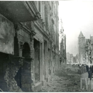 zagruzowana ulica, jej środkiem idą lidzie, po jej obu stronach wypalone kamienice