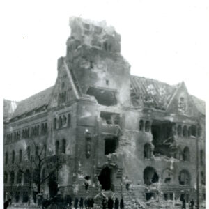 zniszczony i wypalony duży narożny budynek, przed nim ludzie