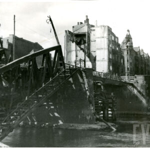 zniszczony i prowizorycznie zabezpieczony most nad rzeką, po nim idą ludzie, w głębi wysokie kamienice, część z nich zniszczona