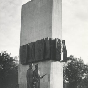prosta, pionowa, gładka, wysoka, prostokątna bryła, w połowie jej wysokości wiszą ciemne tablice z płaskorzeźbami przedstawiającymi sylwetki ludzi, przed bryłą stoi brązowa rzeźba dwóch żołnierzy w wojskowych płaszczach i rogatywkach na głowach, w dłoniach trzymają broń, jeden karabin, drugi opuszczoną szablę
