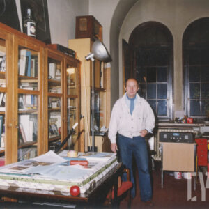 pomieszczenie archiwum z przeszklonymi szafami, regałami, stołem i biurkiem wypełnione książkami, plakatami i zdjęciami, w głębi dwa duże okna, na tle których stoi mężczyzna w jasnej kurtce i dżinsach