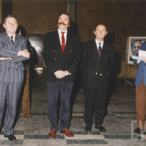 czterech mężczyzn w garniturach stoi w pomieszczeniu z marmurową posadzką, w tle stelaże wystawy fotografii