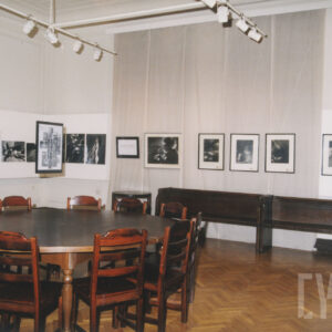 pomieszczenie o białych ścianach, częściowo osłoniętych płótnem, a których wiszą fotografie, na pierwszym planie okrągły stół otoczony krzesłami