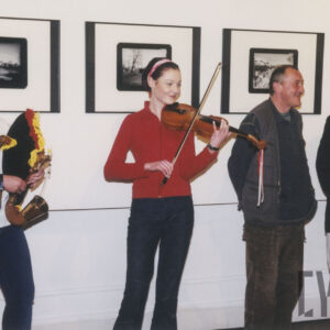 na tle wiszących na ścianie fotografii stoją cztery osoby, dwie kobiety, pierwsza od lewej gra na dudach, druga na skrzypcach, i dwaj rozmawiający mężczyźni