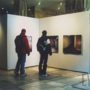 dwoje ludzi stoi przed ścianką działową we wnętrzu dużego pomieszczenia i ogląda zawieszone na niej zdjęcia