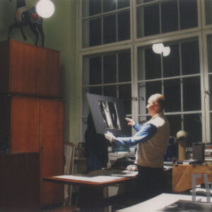 w ciemnym i zagraconym pokoju z dużą liczbą różnych mebli i wielkim oknem stoi mężczyzna oglądający trzymaną w rękach fotografię naklejoną na karton