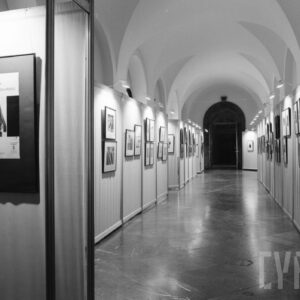 długi, łukowo sklepiony korytarz z drzwiami na końcu, wzdłuż ścian na naciągniętych na ramy płótnach po obu stronach wiszą oprawione fotografie, na pierwszym planie plakat wystawy