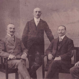 pięciu mężczyzn w różnym wieku w eleganckich garniturach z początku XX wieku; trzech siedzi na krzesłach, dwóch stoi za nimi; wszyscy mają krótko przystrzyżone włosy i wąsy, pozują na tle dekoracyjnej ściany