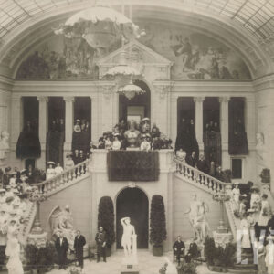 eleganccy, odświętnie ubrani ludzie pozujący do fotografii stojąc w dużym holu z monumentalnymi schodami, wysokim przeszklonym sklepieniem i dekoracjami