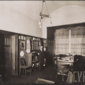 mężczyzna w średnim wieku ubrany w garnitur, pozujący do fotografii w dużym pomieszczeniu, w którym stoją m.in. biurko, krzesła, na ścianach wiszą obrazy