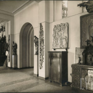 przestronna sala z eksponatami sakralnymi i rzeźbami; widoczne arkadowe przejście, stiukowe pilastry z ornamentem roślinnym, drewniane rzeźby na postumentach, na ścianach płaskorzeźba oraz obrazy, widoczne też zabytkowa szafka i ołtarzyk z krucyfiksami, świecznikami i monstrancją, pod sufitem żyrandol z świecami