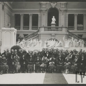 eleganccy, odświętnie ubrani ludzie pozujący do fotografii stojąc w dużym holu z monumentalnymi schodami, wysokim przeszklonym sklepieniem i dekoracjami