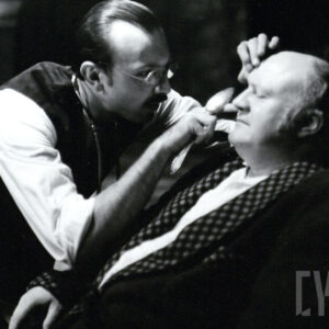 mężczyzna w średnim wieku w okularach nachyla się i przygląda się starszemu, siedzącemu mężczyźnie ubranemu w szlafrok