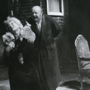 starsza kobieta w sukni z owiniętym wokół szyi szalem imitującym futro z lisa, w tle starszy mężczyzna w szlafroku