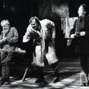 czterech mężczyzn, najmłodszy z nich siedzi przy stole i ma zdziwioną minę, trzech starszych stoi, jeden opiera się o stół