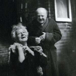 starsza kobieta w sukni z owiniętym wokół szyi szalem imitującym futro z lisa, w tle starszy mężczyzna w szlafroku