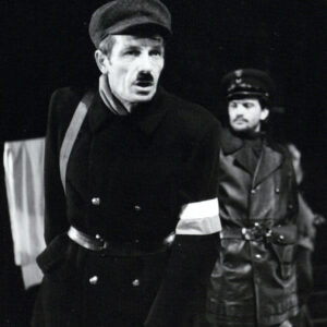 dwóch mężczyzn w średnim wieku, obaj mają wąsy, mężczyzna na pierwszym planie ubrany jest w czapkę, długi płaszcz z biało-czerwoną opaską na ręku, stojący za nim mężczyzna ubrany jest w strój milicjanta 