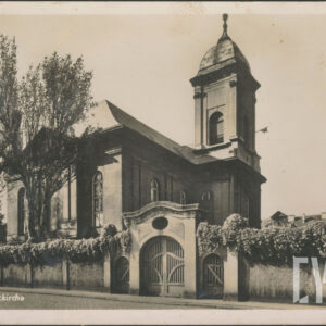 na pierwszym planie murowane ogrodzenie w górnej części porośnięte roślinnością z okazałą bramą i furtkami po obu jej stronach, za grodzeniem budynek sakralny ze spadzistym dachem i wieżą
