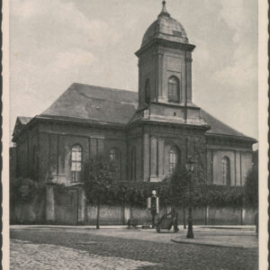 budynek sakralny z przylegającą do niego pośrodku wieżą, otoczony murowanym ogrodzeniem w górnej części porośniętym roślinnością, na pierwszym planie brukowana ulica, pośrodku niej mężczyzna pcha dwukołowy wózek