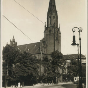 na pierwszym planie ulica, w głębi drzewa, za nimi neogotycka ceglana budowla sakralna z wysoką wieżą, z prawej, przy ulicy latarnia uliczna