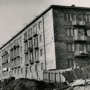 nieotynkowany blok z rzędami okien i balkonami w budowie, po prawej walący się drewniany płot