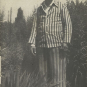 starszy mężczyzna w pasiastej piżamie, stoi na tle krzewów i uśmiecha się pozując do fotografii