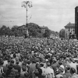 bardzo duża liczba ludzi zgromadzonych na placu, w głębi okazałe budynki, pośrodku latarnia, na jej cokole stoi kilka osób