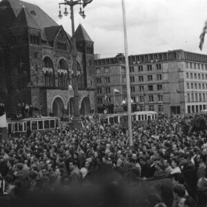 bardzo duża liczba ludzi zgromadzonych na ulicy, w głębi okazałe budynki, wśród tłumu tramwaje, na ich dachach stoją grupki ludzi