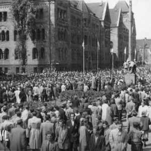 bardzo duża liczba ludzi zgromadzonych na ulicy, pośrodku tłumu stoją tramwaje i ciężarówki, na ich dachach stoją grupy ludzi, w głębi okazałe budynki 