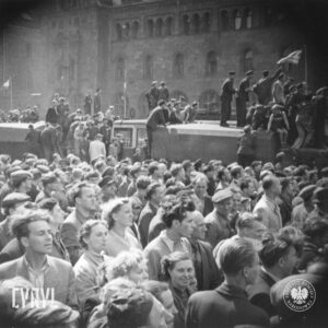 bardzo duża grupa ludzi zgromadzona na ulicy, wśród tłumu dwie ciężarówki, na ich dachach stoją ludzie, część osób wchodzi na ich dachy, w głębi fragment okazałego budynku