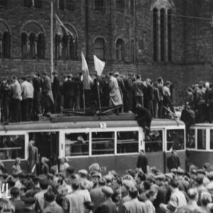 bardzo duża grupa ludzi zgromadzona przed monumentalnym budynkiem, pośrodku tramwaj, na jego dachu stoją ludzie, przy pantografie dwie flagi