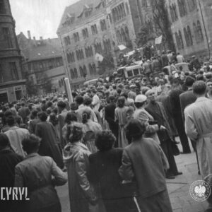 bardzo duża grupa ludzi zgromadzona na ulicy, pośrodku samochód ciężarowy z megafonami, na nim stoją ludzie, część trzyma flagi, w głębi monumentalny budynek
