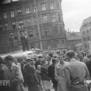 bardzo duża grupa ludzi zgromadzona na skrzyżowaniu ulic, z lewej wśród tłumu stoi autobus, na jego dachu stoi trzech mężczyzn, w głębi okazała kamienica z kilku jej okien wyglądają ludzie
