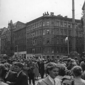 bardzo duża grupa ludzi zgromadzona na skrzyżowaniu ulic, w głębi okazałe kamienice, na dachu kamienicy narożnej stoi kilka osób