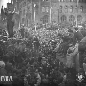 duże zgromadzenie ludzi na ulicy, wśród tłumu ciężarówka z siedzącymi na pace mężczyznami w mundurach milicyjnych, na ciężarówkę wchodzi kilku mężczyzn, w głębi okazałe budynki