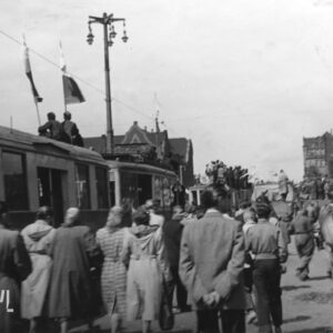 stojące w jednym rzędzie tramwaje, na dachach kilku z nich stoją lub siedzą ludzie, ulicą idą grupy ludzi