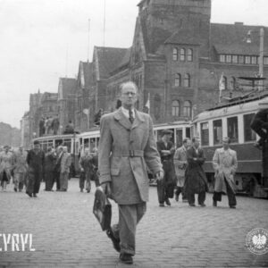 stojące w jednym rzędzie tramwaje, na dachach kilku z nich stoją lub siedzą ludzie, ulicą idą grupy ludzi, na pierwszym planie idzie mężczyzna w płaszczu, w ręce trzyma teczkę, w głębi okazałe budynki