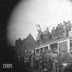 duża grupa ludzi stojąca na dachu tramwaju oraz wokół niego, w głębi okazały budynek z ozdobnym szczytem