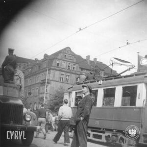 stojący tramwaj na ulicy, na jego dachu siedzi dwóch mężczyzn, ulicą idą grupy ludzi, z lewej fragment szoferki ciężarówki, w głębi okazały budynek