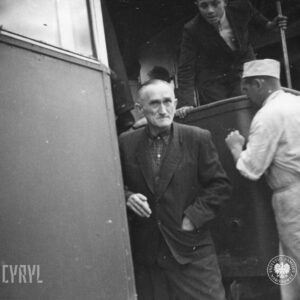 fragment dwóch połączonych ze sobą wagonów tramwajowych, przy nim starszy mężczyzna, z prawej mężczyzna w białej koszuli i fartuchu oraz furażerce na głowie, na otwartym pomoście wagonu tramwajowego stoi mężczyzna