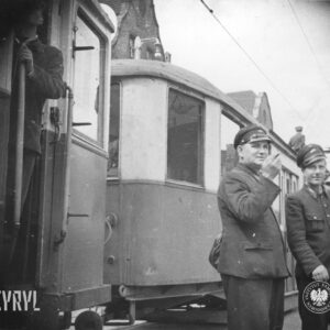 dwóch mężczyzn w mundurach stoi przy dwóch, połączonych ze sobą wagonach tramwajowych
