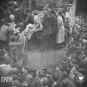 bardzo duża liczba ludzi zgromadzonych na ulicy, pośrodku tłumu ciężarówka, na jej pace stoją lub siedzą mężczyźni i chłopcy