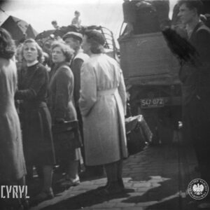 kilka kobiet i mężczyzn zgromadzonych wokół samochodów ciężarowych, z tego dwie kobiety trzymają w rękach teczki